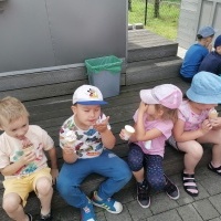 Dzieci siedzą na ławeczce. Jedzą lody. W tle lodziarnia.