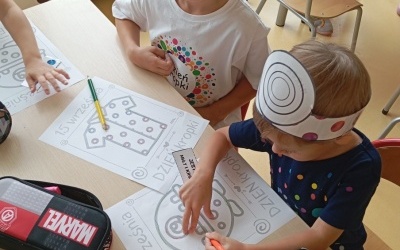 Dzieci siedzą przy stolikach i kreślą po śladzie obrazek. Na głowach mają opaski z dużą kropką