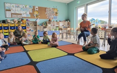 Dzieci siedzą na dywanie. Pani bibliotekarka gra na ukulele. 