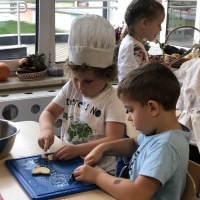 Dwoje chłopców kroi przy stoliku owoce na desce kuchennej. W tle inne dzieci przygotowujące sałatkę.