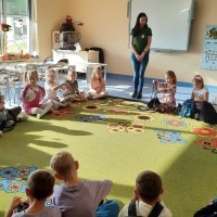 Dzieci siedzące w kole na dywanie. W kole stoi Pani Bibliotekarka. W tle tablice, stoliczki i biurko nauczyciela.