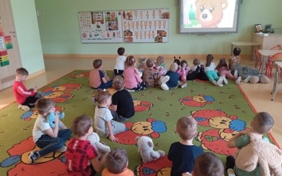Dzieci siedzą na dywanie trzymając swoje pluszowe Misie i oglądają bajkę o Misiach  