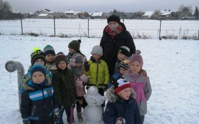 Grupa dzieci wraz z Panią stoją  na placu przedszkolnym w kole a w środku bałwanek zbudowany przez dzieci. Zimowa sceneria, w tle ogrodzenie i pola.                                    