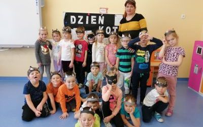 Dzieci wraz z panią pozujący do zdjęcia grupowego na tle tablicy z napisem Dzień KOTA. Dzieci mają na głowach opaski z uszkami kota. 