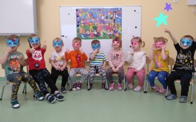 Dzieci siedzą na krzesełkach, na oczach mają założone kolorowe maski. W tle tablica magnetyczna.