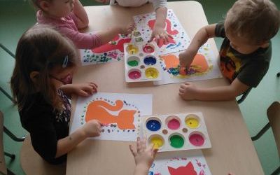 Dzieci siedzą przy stoliku i malują farbkami obrazek kota.