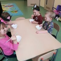Dzieci siedzą przy stoliku i piją mleko z białych miseczek. W tle kolorowy dywan.
