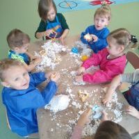 Dzieci ubrane w kolorowe fartuszki siedzą przy stole i bawią się sztucznym śniegiem.