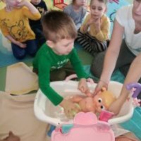 Chłopczyk w zielonej bluzce wraz z panią nauczycielką myją lalkę, która znajduje się w wannie wypełnionej wodą. 