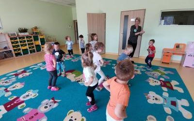 Grupa dzieci wraz z panią tańczą na dywanie. W tle drzwi oraz kolorowe szafki na zabawki. 