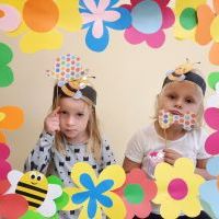 Dwie dziewczynki w ramce z kolorowych kwiatów. Na głowach mają opaski z pszczółkami, w rękach kolorowe rekwizyty.