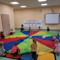 Grupa dzieci wraz z nauczycielką bawi się kolorową chustą i balonami. W tle tablica magnetyczna z kolorowym napisem 