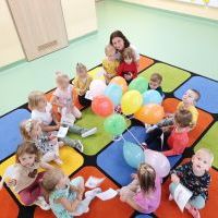 Grupa dzieci siedząca w kole na dywanie z nauczycielką. W rękach dzieci chorągiewki. W środku koła wazon z kolorowymi balonami.