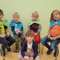 Grupa chłopców siedzi na krzesełkach. Na szyi mają kolorowe muszki z papieru. Przed chłopcami na podłodze siedzi dziewczynka ubrana w granatowy sweterek. 