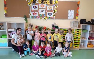 Dzieci ustawione przed półkami. Z lewej strony nauczyciel. Za nimi jesienny kącik z warzywami oraz tablice korkowe.
