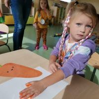 Dziewczynka w pastelowych warkoczykach wykonuje pracę plastyczną odbijając dłoń maczaną w pomarańczowej farbie. W tle dywan, bawiące się dzieci, nogi Pani, szafki.   