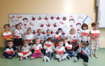 Dzieci ustawione w dwóch rzędach przed tablicą. W rękach trzymają biało - czerwone serduszka. Na tablicy napis Kocham Cię Polsko oraz obrazki flagi polskiej.