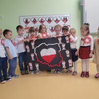 Grupa dzieci ustawiona w jednym rzędzie trzyma dużą mapę Polski wykonaną z kolorowej bibuły. 