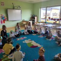 Grupa dzieci siedzi na kolorowym dywanie. Dzieci mają na głowie opaski z wizerunkiem motylka. W tle duże okno i tablica interaktywna.