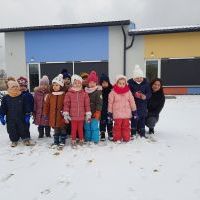 Grupa dzieci z nauczycielem na placu zabaw w zimowy dzień. Za nimi budynek przedszkola.