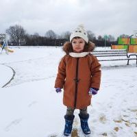 Chłopiec w zimowym stroju stoi na placu zabaw. W tle ławeczka, ogrodzenie boiska oraz drzewa.