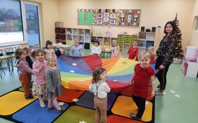 Grupa dzieci wraz z nauczycielem stoi w kole, trzymają w rękach kolorową chustę animacyjną. W tle półki z zabawkami oraz tablice korkowe z dekoracjami i obrazkami.