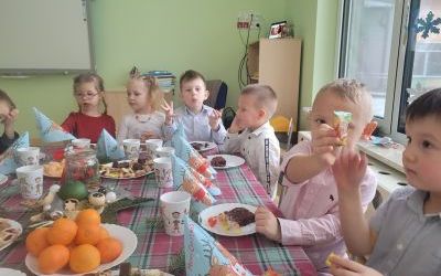 Dzieci siedzą przy świątecznym stole. Na stole znajdują się słodycze, mandarynki i ozdoby świąteczne. 