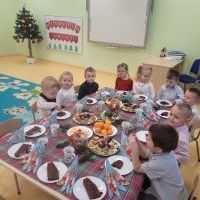 Dzieci siedzą przy świątecznym stole. Na stole znajdują się słodycze, mandarynki i ozdoby świąteczne. W tle stoi choinka.