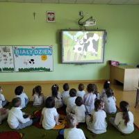 Dzieci siedzą na kolorowym dywanie, patrzą na ekran tablicy multimedialnej      i oglądają film edukacyjny dla dzieci pt. „Skąd się bierze mleko”. W tle tablica magnetyczna z obrazkami.