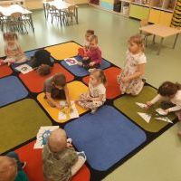 Dzieci siedzące na dywanie układają puzzle przedstawiające bakterie. W tle stoliki z krzesełkami oraz półki z zabawkami.