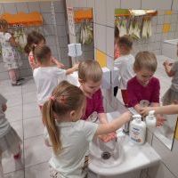Łazienka przedszkolna. Cztery dziewczynki myją ręce. Z lewej strony dziewczynka idąca w stronę ręczników. W tle dziewczynka wycierające ręce.