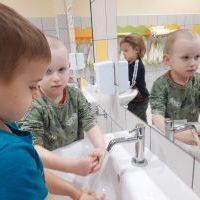 Łazienka przedszkolna. Na pierwszym planie dwaj chłopcy myjący ręce. W tle chłopiec oraz wieszaczki z ręcznikami.
