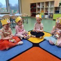 Cztery dziewczynki w koronach siedzą na dywanie i oglądają upominki z okazji Dnia Kobiet. Za nimi półki z zabawkami i stoliki z krzesełkami.
