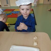 Chłopiec w czapce piekarza siedzi przy stoliku i lepi z masy solnej pieczywo. W tle bawiące się dzieci.