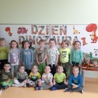 Grupka dzieci ustawiona w dwóch rzędach przed tablica magnetyczną, na której widnieje napis dzień dinozaura. Z prawej strony dzieci stoi stoliczek z eksponatami dinozaurów