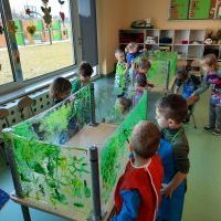 Dzieci ubrane w fartuszki malują zieloną farbą na folii. W tle okna wychodzące na plac zabaw oraz półki z zabawkami.