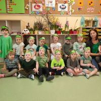 Grupa dzieci ustawiona w dwóch rzędach. Z prawej strony nauczyciel trzymający na kolanach chłopca. Dzieci mają na głowach zielone opaski. Za nimi kącik przyrodniczy z wiosennymi kwiatami oraz tablice korkowe z obrazkami.