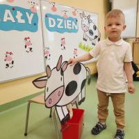 Chłopiec ubrany na biało stoi przy dużej sylwecie krowy wyciętej z kartonu. Za nim w tle tablica magnetyczna z napisem Biały Dzień i sylwetami krówek.