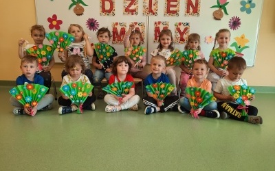 Dzieci z grupy Pszczółki siedzą przy tablicy. W rękach trzymają papierowe bukieciki. W tle napis Dzień Mamy.