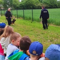 Policjanci prowadzą psa policyjnego. Dzieci stoją na boisku. W tle płot i domy.