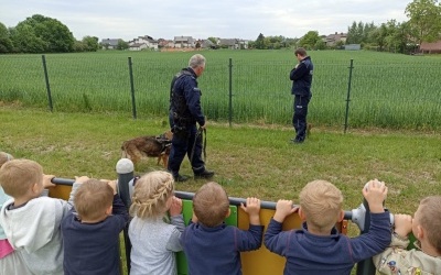 Policjant prezentuje komendy, które wykonuje pies policyjny. Dzieci stoją na boisku przedszkolnym. W tle ogrodzenie, domy i drzewa. 