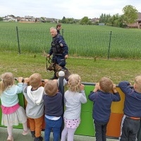 Policjant prezentuje komendy, które wykonuje pies policyjny. Dzieci stoją na boisku przedszkolnym. W tle ogrodzenie, domy i drzewa. 
