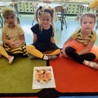Trzy dziewczynki siedzące na dywanie. Przed nimi obrazek przedstawiający ul i pszczółki. W tle stoliki z krzesłami. 