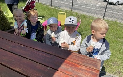 Grupa dzieci siedzi na ławeczce i zjada loda. W tle ogrodzenie, samochody i budynek. 