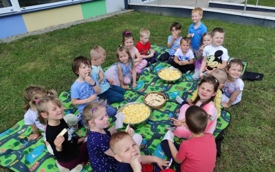 Dzieci siedzą na kocu, na podwórku przedszkolnym, uśmiechają się i jedzą chrupki i wafelki.