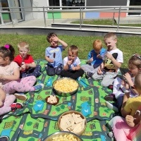 Dzieci siedzą na kocu, na podwórku przedszkolnym częstują się: chrupki, wafelki, lodami.