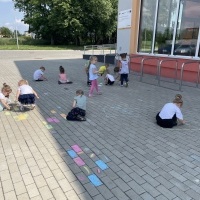 Grupa dzieci maluje kredą na kostce brukowej na parkingu przedszkolnym. 