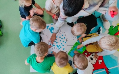 Dzieci z grupy Elfy siedzą wokół dużej kartki białego papieru z namalowana Kicią Kocią wraz z Panią i przyklejają małe karteczki w kolorze niebieskim na kontur Kota. Rzut z góry.. 