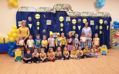 Dzieci w strojach niebiesko-żółtych ustawione w dwóch rzędach pozują do grupowego zdjęcia. Z prawej i lewej strony Panie. Za nimi na niebieskim tle obrazki, dekoracje z bajki 