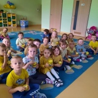 Dzieci ubrane na żółto - niebiesko siedzą na dywanie. W rękach trzymają kubeczki z popcornem. W tle drewniane drzwi, półki z szufladkami dzieci.